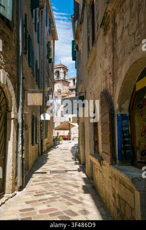 Schmale Kopfsteinpflasterstraße in historischer Stadt mit Steinbauten, die grüne Fensterläden und einen Kirchturm unter klarem blauen Himmel aufweisen. Stockfoto