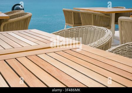 Holztische und Korbstühle, die auf einer Terrasse mit Blick auf das Meer arrangiert sind. Die Szene vermittelt eine entspannte Atmosphäre für das Essen im Freien. Stockfoto