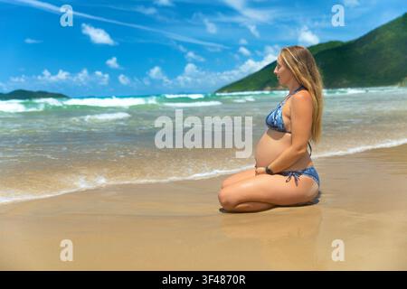 Neun Monate schwangere Frau im dritten Trimester meditiert und entspannt sich an einem tropischen Meeresstrand. Gesunder pränataler Lebensstil, Reisen im Sommerurlaub Stockfoto