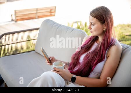 Junge Frau mit langen rosa Haaren entspannt sich auf einem Terrassensofa, hält ein Smartphone und eine Tasse Kaffee bei hellem Tageslicht und genießt ruhige Freizeit und fr Stockfoto