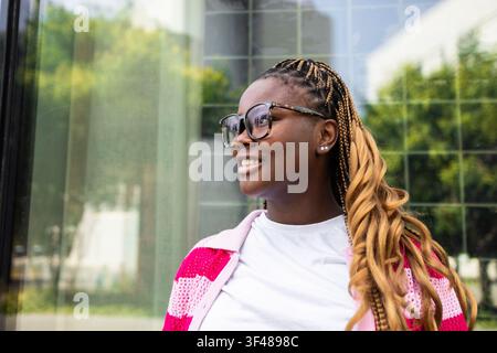Seitenansicht einer lächelnden schwarzen Frau mit langen blonden Zöpfen und einer Brille, die einen pink gestreiften Pullover und ein weißes Oberteil trägt und neben einem modernen Glas steht Stockfoto