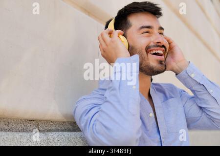 Nahaufnahme eines lächelnden jungen Mannes mit gelben Kopfhörern, der beim Musikhören im Freien neben einer Steinmauer lacht und Entspannung, Freude und ein Stockfoto