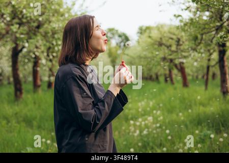Seitenansicht einer Frau, die Löwenzahnsamen in einer üppigen Frühlingswiese in einem Obstgarten bläst, umgeben von grünen Bäumen und sanftem Sonnenlicht, die Natur, Ruhe, ein feiert Stockfoto