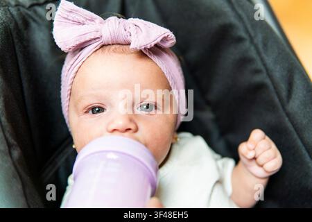 Nahaufnahme-Porträt eines Baby-Mädchens mit einem rosa Schleifen-Stirnband, das aus einer lila Flasche trinkt, während es in einem dunklen Kinderwagen-Sitz ruht, weiches natürliches Licht. Stockfoto