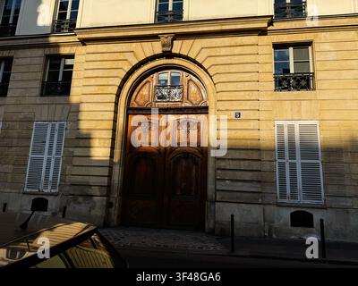 Sonnige verzierte Holztür in einem Kalkstein-Haussmann-Gebäude mit weißen Fensterläden und warmen Schatten. Stockfoto