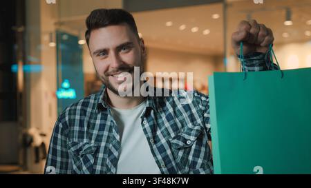 Glücklicher, optimistischer kaukasischer Kundenmann hält eine Einkaufstasche, zeigt lächelnd, posiert als Kunde mit Kaufpaket, Geschenk, Lächeln, Mann genießt die Verkäufe Stockfoto