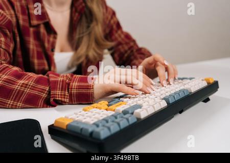 Frau in rotem kariertem Hemd tippt auf einer benutzerdefinierten mechanischen Tastatur. Schwache Tiefenschärfe lenkt den Fokus auf gepflegte Hände und bunte Tasten. Moderner Arbeitsbereich Stockfoto
