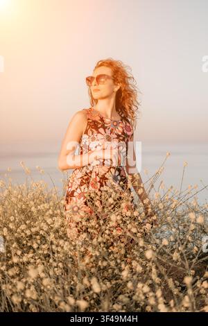 Frau Blumen Sonnenuntergang, rothaarige Frau in Blumenkleid steht zwischen Wildblumen am Strand während der goldenen Stunde für einen Sommer-Lebensstil Stockfoto