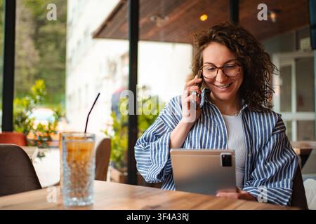 Frau telefoniert am Smartphone und verwendet ein Digitaltablett im Café Stockfoto