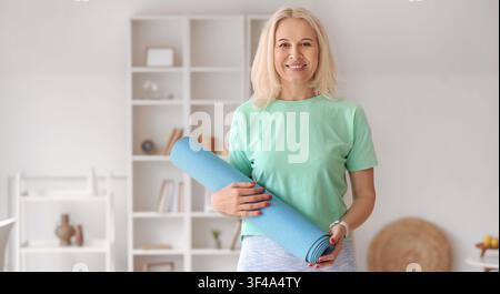 Lächelnde reife Frau hält eine blaue Yogamatte, bereit für Fitness und einen gesunden Lebensstil zu Hause Stockfoto