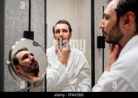 Mann in einem weißen Bademantel, der sein Spiegelbild im Badezimmerspiegel betrachtet, seinen Bart und Hals berührt und sich auf persönliche Pflege und Selbstfürsorge konzentriert Stockfoto