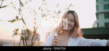 Junge Frau lächelt, hält ein Glas und genießt einen glücklichen Lebensstil bei Sonnenuntergang Stockfoto