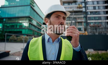 Moving Shot Kaukasischer Mann Bauingenieur in Hardhat Talk Remote Radio Outdoor Urban Building Architekt Bauunternehmer mit Helm Sicherheitsweste Stockfoto