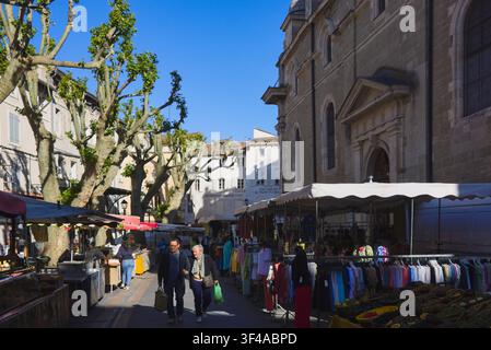 Besucher erkunden belebte Marktstände im Freien, umgeben von Platanen und historischen Gebäuden in L'Isle-sur-la-Sorgue, Frankreich. Stockfoto