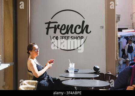 Eine Frau, die an einem Tisch auf einer Außenterrasse sitzt und ein Smartphone benutzt, außerhalb eines französischen Cafés in L'Isle-sur-la-Sorgue, Provence, Frankreich. Stockfoto