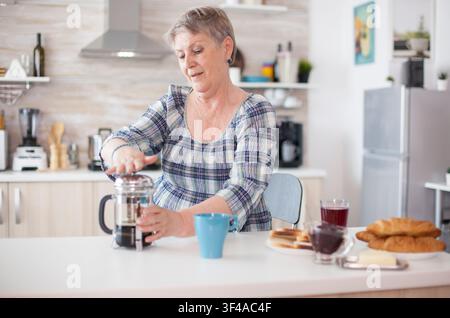 Frischer Kaffee wird aus einer French Press eingegossen, während eine ältere Frau ihr Frühstück genießt, was Koffein, häuslichen Komfort und die Zubereitung von Morgengetränken zeigt. Stockfoto