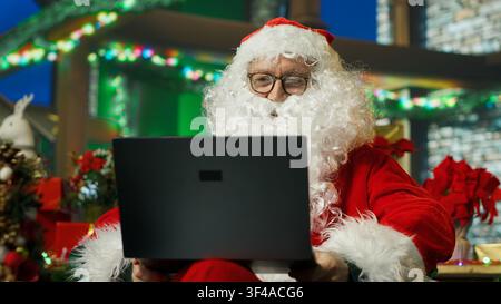 Weihnachtsmann macht Internet-Video mit Kindern auf Laptop aus dem gemütlichen Weihnachtsset. Mann, der Kinder in Notebook-Videokonferenz anruft, nachdem er sich ihre Wunschlisten angesehen hat, Kamera A Stockfoto