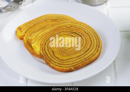 Nahaufnahme eines gerollten indonesischen Lapis-Legit-Kuchens auf einem weißen Teller. Traditionelles geschichtetes Dessert mit goldener Farbe und sauberem Food-Styling. Stockfoto