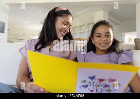 Afroamerikanische Mutter und Tochter tragen malvenfarbene Bluse, sitzen auf dem Sofa und halten eine große gelbe Karte Stockfoto