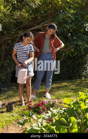 Afrikanische Mutter beobachtet, wie das Kind rote rosafarbene Blumen im Hof mit einer blassrosa Gießkanne gießt Stockfoto