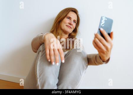 Mittealter Frau mit blondem Haar sitzt auf dem Boden, macht ein Selfie, nutzt ein Smartphone, kommuniziert online Stockfoto