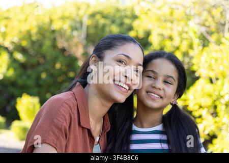 Afroamerikanische Mutter und ihre jugendliche Tochter lehnen sich in einem sonnendurchfluteten Garten an und lächeln, während sie ein gestreiftes T-Shirt tragen Stockfoto