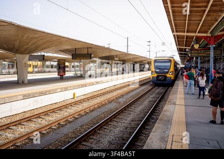 Porto, 17. Juni 2022: Passagiere stehen auf einem belebten Bahnsteig und warten auf einen herannahenden gelben Zug. Stockfoto