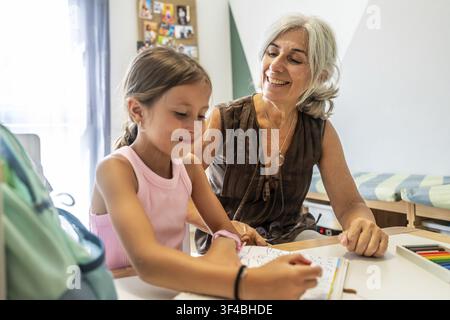 Großmutter sitzt an einem Tisch, lächelt und beaufsichtigt ihre Enkelin, die in einem Buch malt, und fördert so Lernen, Bindung und Familienzusammenhalt in einem Stockfoto