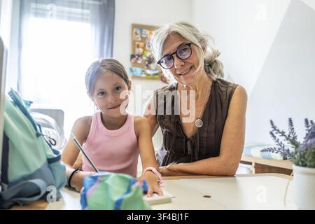 Großmutter und Enkelin teilen einen Bindungsmoment, sitzen an einem Schreibtisch und lächeln in die Kamera, während sie sich mit Lernaktivitäten und Stu Stockfoto