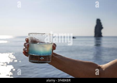 Hand hält ein Glas mit Blue Curacao oder Blue Curacao, Getränk, Gegenlicht, im Hintergrund das Mittelmeer und eine Felsnadel, Äolische oder Äolische Inseln Stockfoto