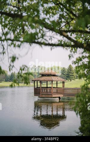 Gartenlaube an einem ruhigen See, eingerahmt von überhängenden Weidenzweigen, Holzpavillon spiegelt sich im stillen Wasser, ferne Kiefern und spätsommerliches Licht Stockfoto