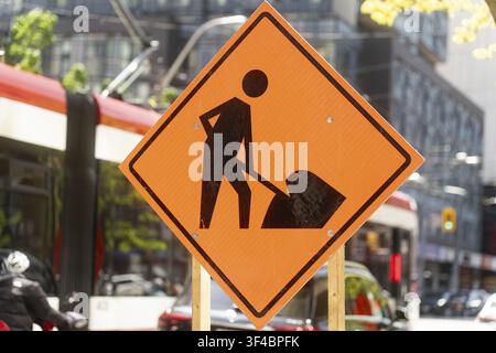 Ein helles orangefarbenes Baustellen-Warnschild mit einem Arbeiter mit einer Schaufel ist im Stadtzentrum von Toronto gut sichtbar angebracht. Im Hintergrund ein Rotes und Stockfoto