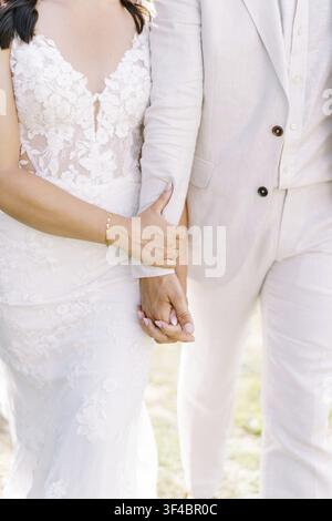 Sonnige Braut und Bräutigam, die sich an den Händen halten, Spitzendetail am Kleid, neutraler Leinenanzug, Gegenlicht im Goldenen Stundenlicht, weicher Fokus-Hintergrund, zärtlicher Griff Stockfoto