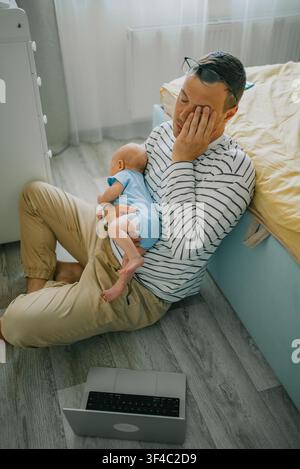 Junger Vater, der emotionalen Burnout und Müdigkeit erlebt, hält sein Baby und reibt sich die Augen, was die Herausforderungen zeigt, die Betreuung der Kinder und die Arbeit von zu Hause aus in Einklang zu bringen Stockfoto