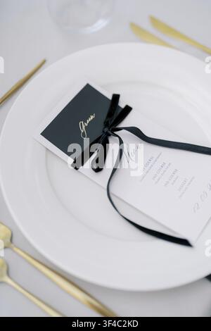 Menü mit schwarzer Schleife auf weißem Teller mit goldenem Besteck. Formelle Tischdekoration im Stil einer Empfangsparty, minimalistisches Porzellan, gebundene Schleife und gedruckte Menükarte Stockfoto