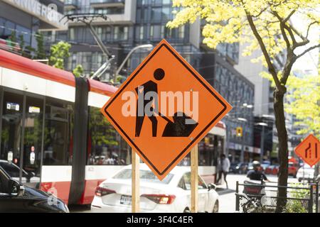 Ein helles orangefarbenes Baustellenschild steht auffällig auf einer belebten Straße in Toronto, mit einer modernen rot-weißen Straßenbahn, Autos und Fußgängern im Stockfoto