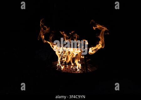 Hell brennendes Holzfeuer in einer runden Metallgrillschale mit glühenden Flammen in der dunklen Nacht. Nahaufnahme des Outdoor-Campingkochaufbaus mit h Stockfoto