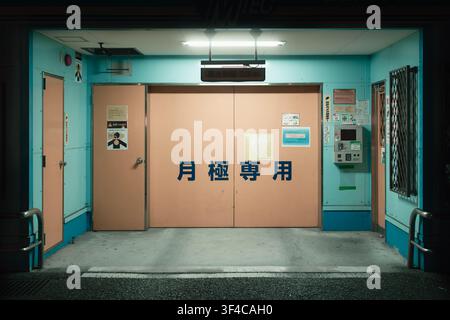Geschlossenes türkisfarbenes Metallschloss eines automatisierten Tiefgarage-Eingangs mit Kanji-Text unter einer Türlampe bei Nacht, Osaka, Japan. Stockfoto