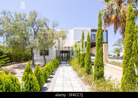 Moderne weiße Luxusvilla mit einem gepflasterten Eingangsweg, der von Zypressen, üppigem grünen Laub und Palmen unter einem strahlend blauen Sommerhimmel gesäumt ist Stockfoto