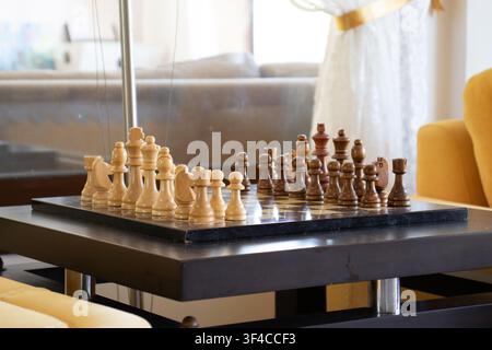 Holzschachbrett mit hellen und dunklen Figuren auf einem modernen Couchtisch in einem hellen, gemütlichen Wohnrauminterieur. Stockfoto