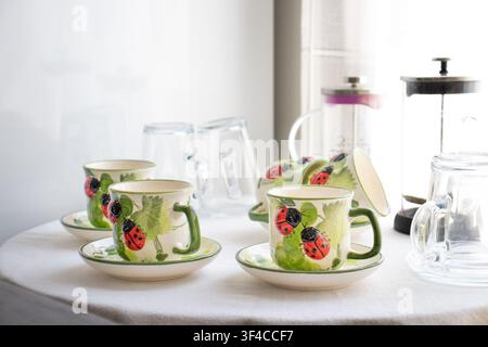 Ein bezauberndes Teeservice mit Keramikbechern und Untertassen, verziert mit handbemalten Marienkäfer- und Blattmotiven, ordentlich auf einer weißen Tischdecke angeordnet Stockfoto