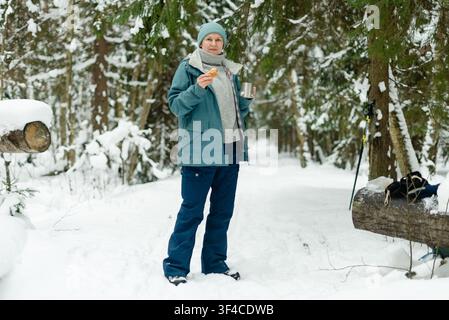 Reife Frau wärmt ihre Hände mit einer dampfenden Tasse und einer Thermoskanne in einem verschneiten Winterwald. Erwachsene Dame nippt an heißem Tee mit sichtbarem Dampf aus der Tasse. Gemütliche Szene von Behaglichkeit und Wärme bei einem kalten Spaziergang im Freien Stockfoto