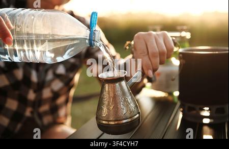 Frau, die Kaffee in Einem Cezve auf Einem tragbaren Campingofen macht und Wasser in Einen türkischen Topf gießt Stockfoto