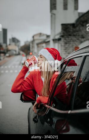Frau trägt roten Pullover und Weihnachtsmannmütze und genießt ein Getränk aus einer Tasse, während sie in einem Auto an der Straße sitzt Stockfoto
