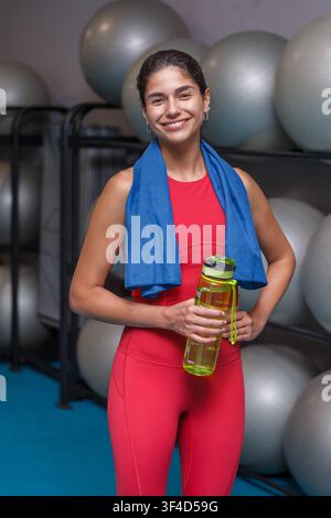Eine Frau steht in einem Fitnessstudio und hält eine Wasserflasche, während ein Handtuch um ihren Hals hängt. Sie trägt Sportkleidung und lächelt. Das Fitnessstudio hat Fitnessbälle in Stockfoto