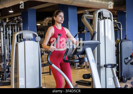 Eine Frau in roter Sportkleidung nutzt ein Crosstrainer zum Training in einem Fitnessstudio. Die Studio-Umgebung bietet verschiedene Trainingsgeräte Stockfoto