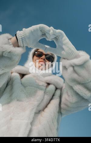 Frau in warmer Kleidung macht ein Herz mit den Händen vor blauem Himmel an einem Wintertag Stockfoto