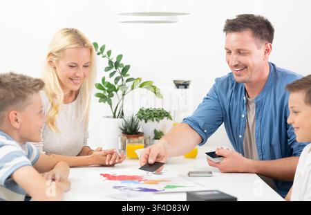 Glückliche Familie spielt gemeinsam ein weltkartengestütztes Lernbrettspiel am Tisch. Vater legt eine Karte auf das Weltkarten-Brettspiel, während die Familie zuschaut und lächelt. Stockfoto