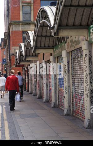 Eine ungezwungene Straßenansicht, die eine Person in einem roten Hemd zeigt, die tagsüber an geschlossenen Ladenfronten entlang einer Stadtstraße vorbeigeht. Stockfoto