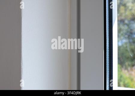 Eine detaillierte Ansicht einer subtil strukturierten weißen Wandfläche mit einer vertikalen Naht und einem dunklen Fensterrahmen, der auf der rechten Seite sichtbar ist Stockfoto